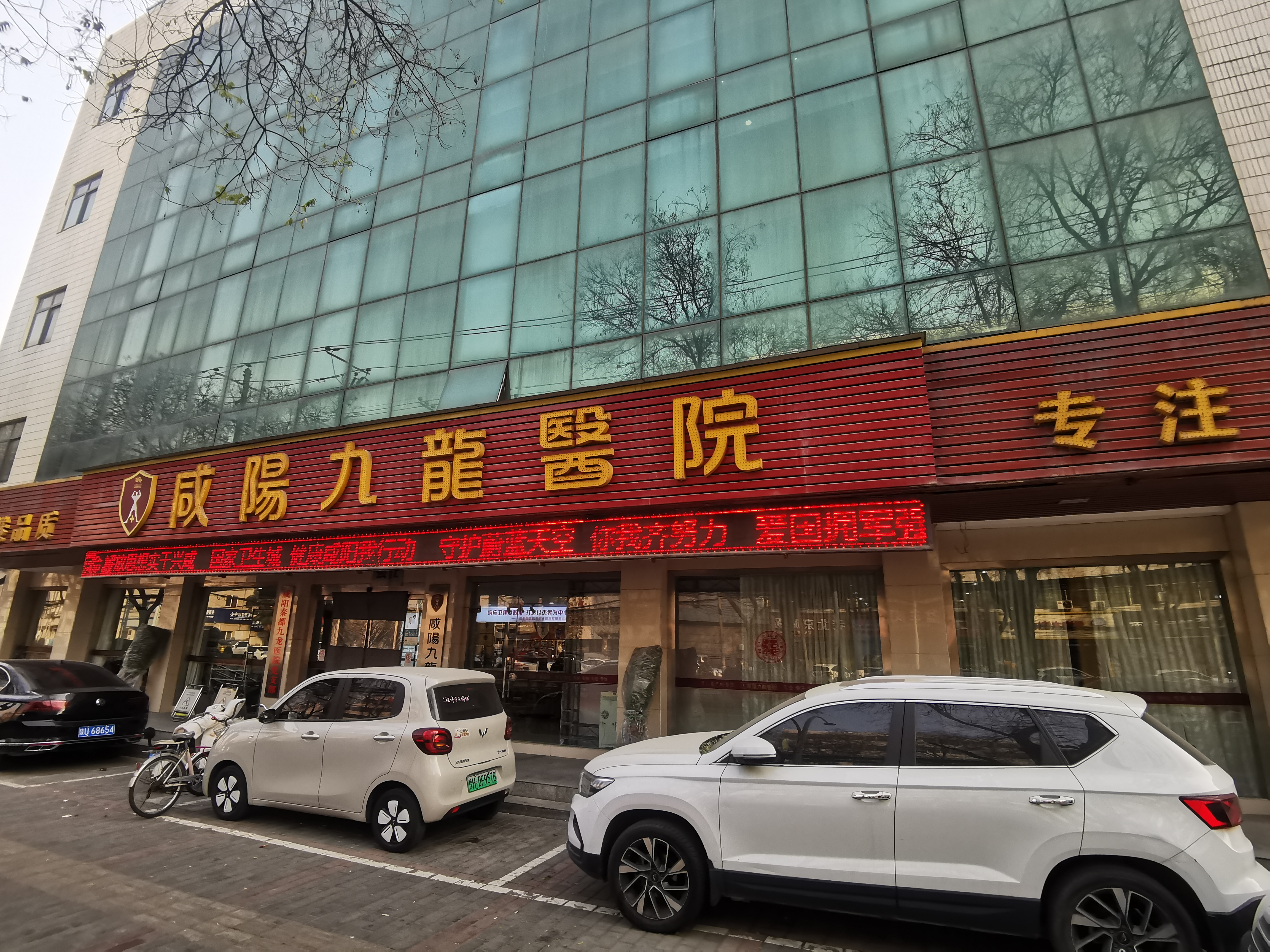 咸阳秦都九龙医院临街外观，电子屏显示公益宣传标语，门前停有多车